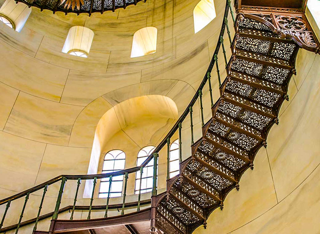 Über zwei halbe Treppen geht es in die 1. Etage und von dort über die wandseitig frei aufgehängte Wendeltreppe auf gußeisernen durchbrochenen Stufen nach oben auf den Turm